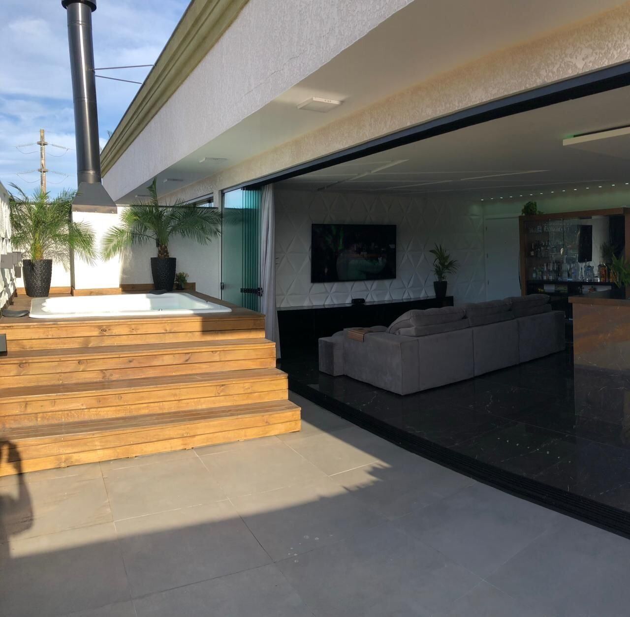 Un área exterior moderna para disfrutar del aire libre, con un jacuzzi, escalones de madera, una zona de estar con sofá y un televisor montado en la pared.