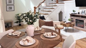 Un coin repas avec une table en bois, dressée pour quatre personnes, entourée de chaises. Un escalier avec des rampes en bois mène à l’étage supérieur.