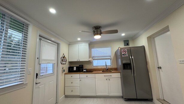Una cocina compacta con armarios blancos, un refrigerador de acero inoxidable, un ventilador de techo y una ventana con persianas.