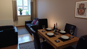 Un comedor con una mesa de madera dispuesta para cuatro personas, rodeada de sillas negras. En el fondo, se ve un sofá y una ventana con cortinas.
