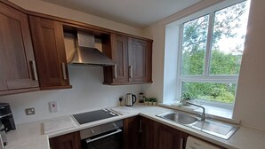Cocina compacta con armarios de madera, horno integrado, lavadora y ventana con vistas a zonas verdes.