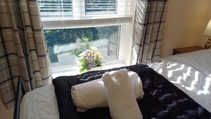 Una habitación de hotel con cama, ventana con cortinas, mesita de noche con lámpara y un ramo de flores en el alféizar.