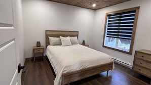 A bedroom with a wooden bed, white walls, a window with blinds, and a wooden floor. - Welcome to your haven of peace, a magnificent chalet nestled in the heart of nature. (Rivière-Rouge)