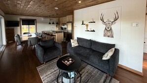 A modern living room with a grey sofa, a coffee table, and a dining area with a kitchen in the background. - Welcome to your haven of peace, a magnificent chalet nestled in the heart of nature. (Rivière-Rouge)