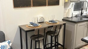 A small kitchen with a dining area, featuring a table with two bar stools, a coffee maker, and a window with blinds.