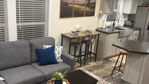 A modern living room with a grey sofa, a black coffee table, and a dining area with a table and chairs. There is a kitchen area with a sink and a refrigerator.