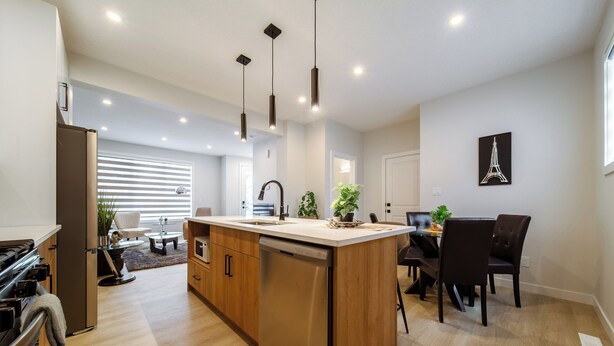 Una cocina moderna con una isla central, armarios de madera, electrodomésticos de acero inoxidable, iluminación colgante y un comedor con mesa y sillas negras.