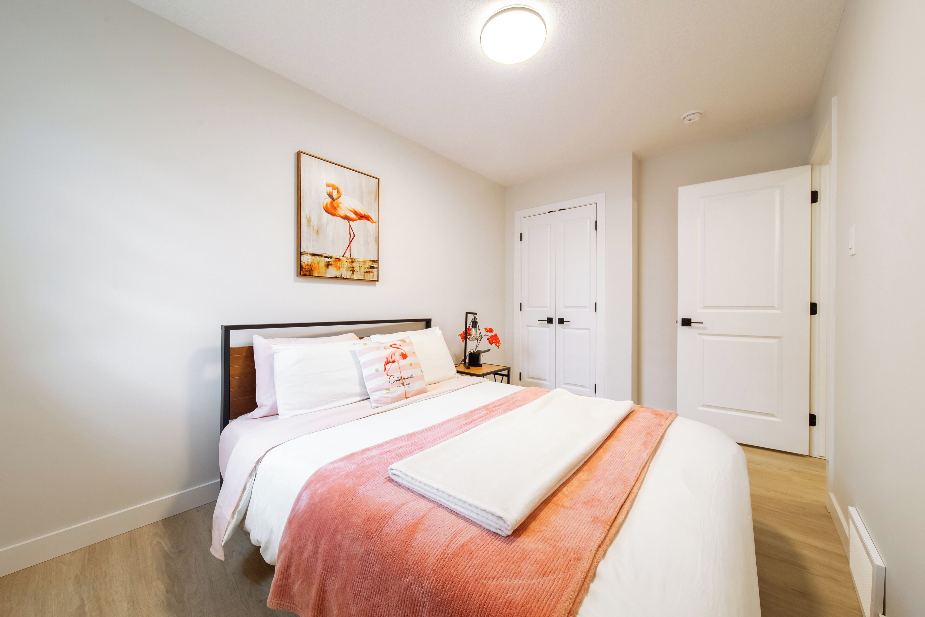 Une chambre à coucher bien rangée, avec un lit, une table de chevet dotée d’une lampe et un tableau encadré accroché au mur.
