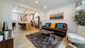 Una sala moderna con una mesa de centro de vidrio, un sofá de cuero marrón y un sillón blanco. Se aprecia una cocina al fondo con un comedor.