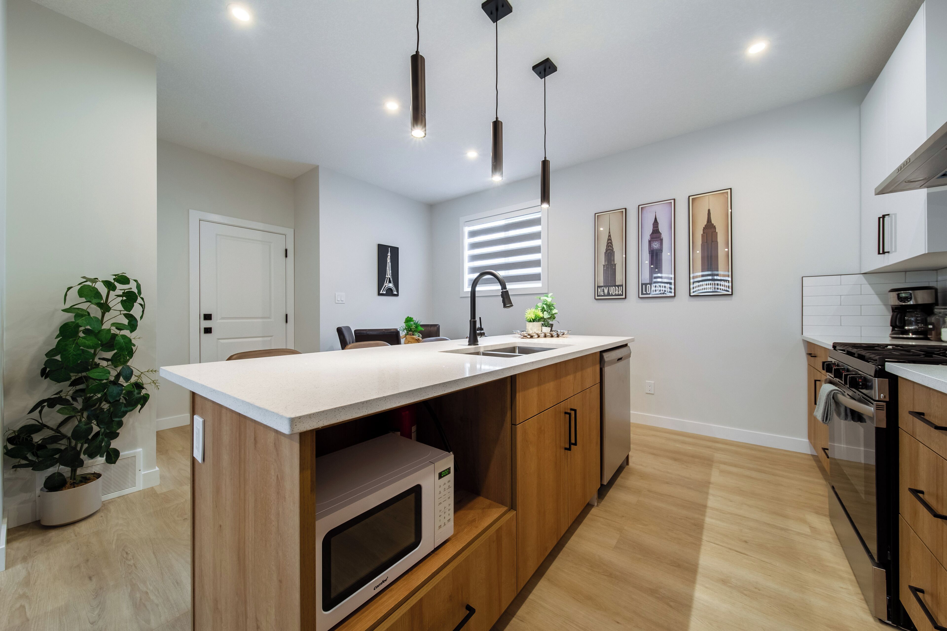 Une cuisine moderne avec des armoires en bois, un plan de travail blanc, un four à micro-ondes encastré et un évier. On y trouve des luminaires suspendus, une plante en pot et des tableaux encadrés au mur.