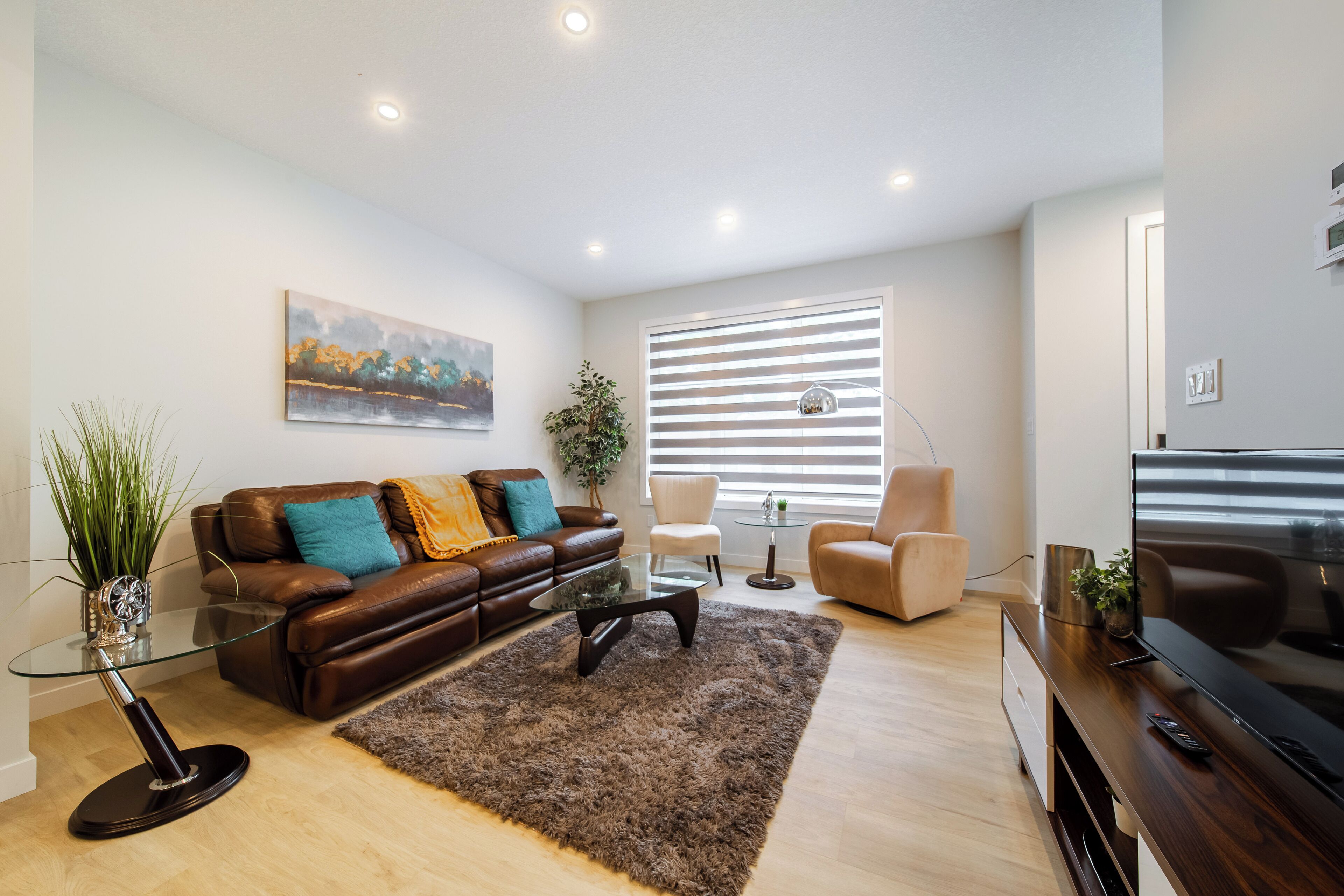 A modern living room with a brown leather sofa, glass coffee table, and a large flat-screen TV. There is a wooden entertainment center, a small side table, and a large window with blinds.