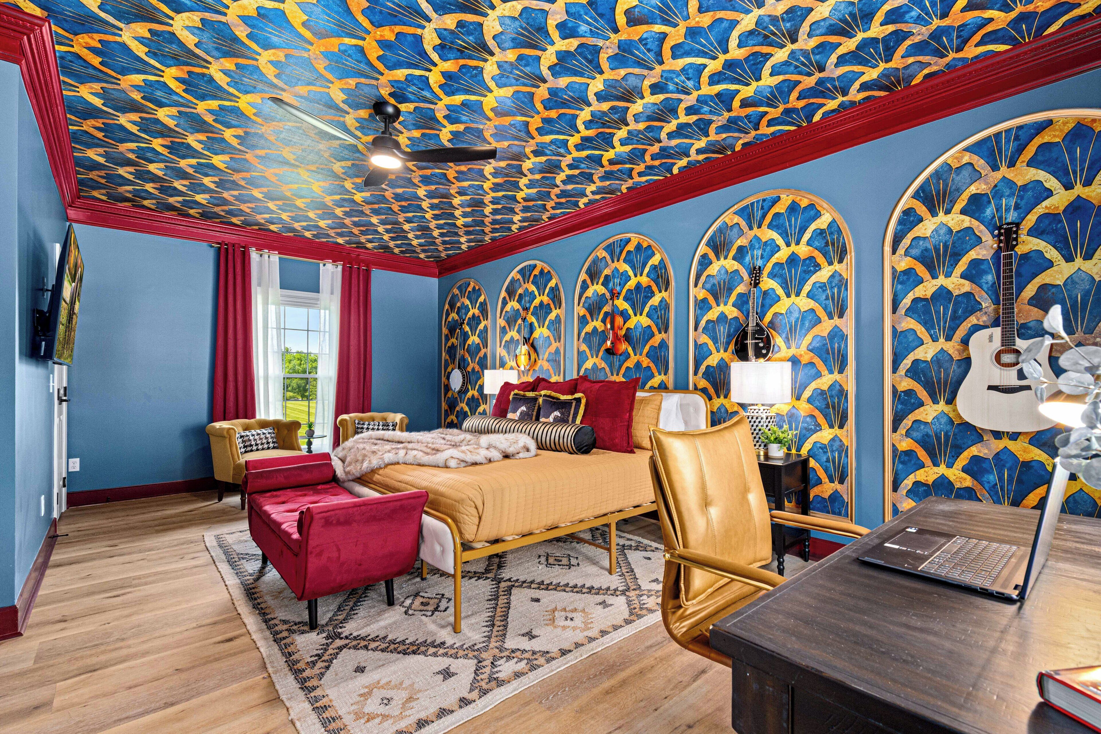 Une chambre à coucher avec un plafond à motifs, un lit avec une literie rouge et dorée, un ordinateur portable sur un bureau et une guitare dans un coin.