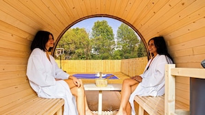 Two people in a sauna with wooden walls and benches, a table with a mat and a bucket, and a view of trees outside.