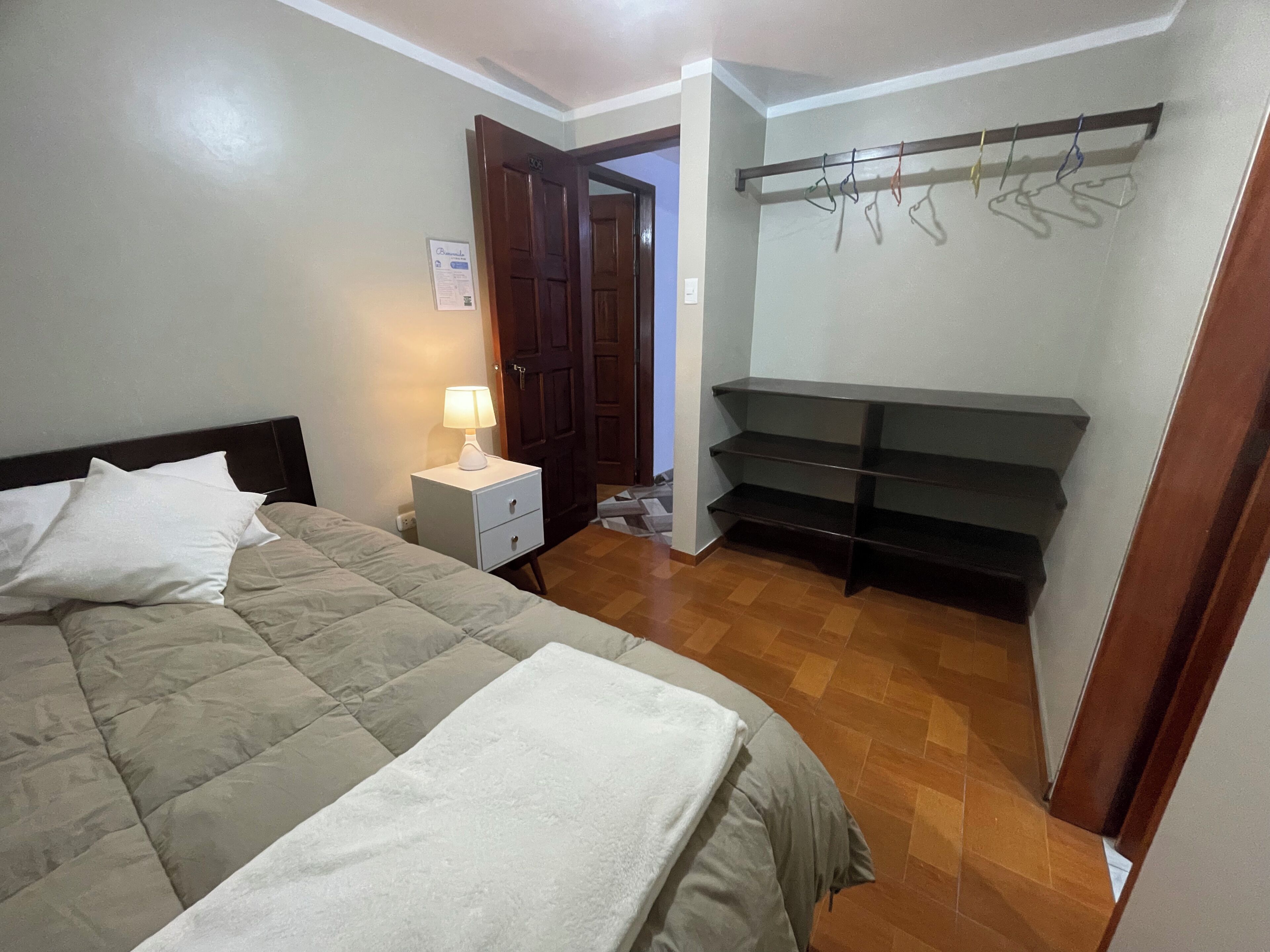 Un dormitori amb un llit, una tauleta de nit amb una làmpada, un armari amb prestatges i una porta que condueix a una altra habitació.