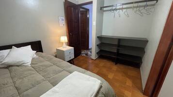 Un dormitori amb un llit, una tauleta de nit amb una làmpada, un armari amb prestatges i una porta que condueix a una altra habitació.