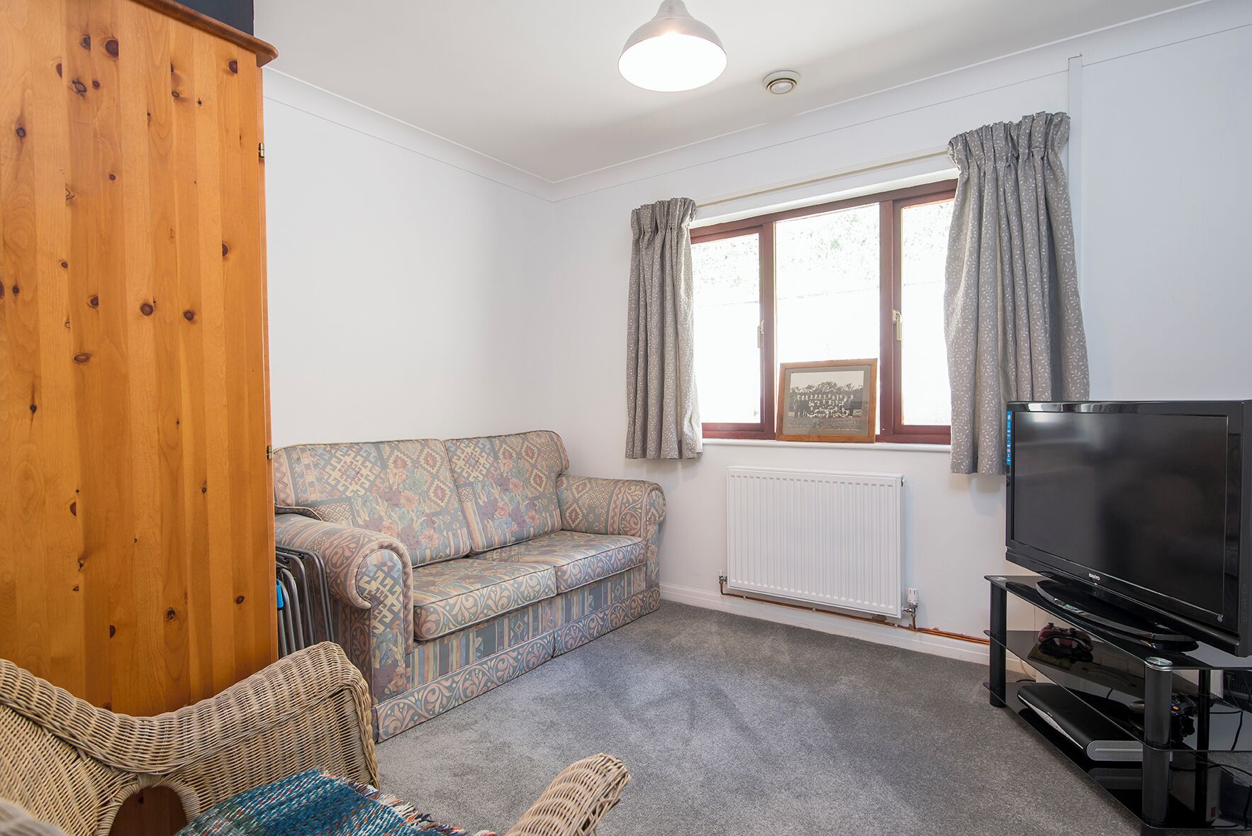 A living room with a patterned sofa, a television on a stand, a radiator, and a window with curtains.
