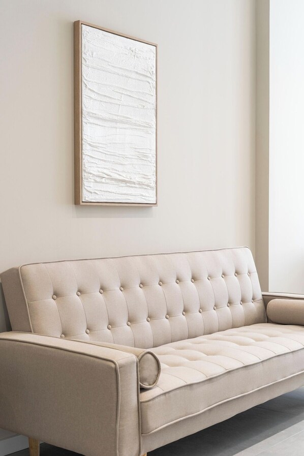 A beige tufted sofa with a wooden frame against a plain wall, positioned in a room with a large window. - VLC HOST - Sumsi B (Valencia)