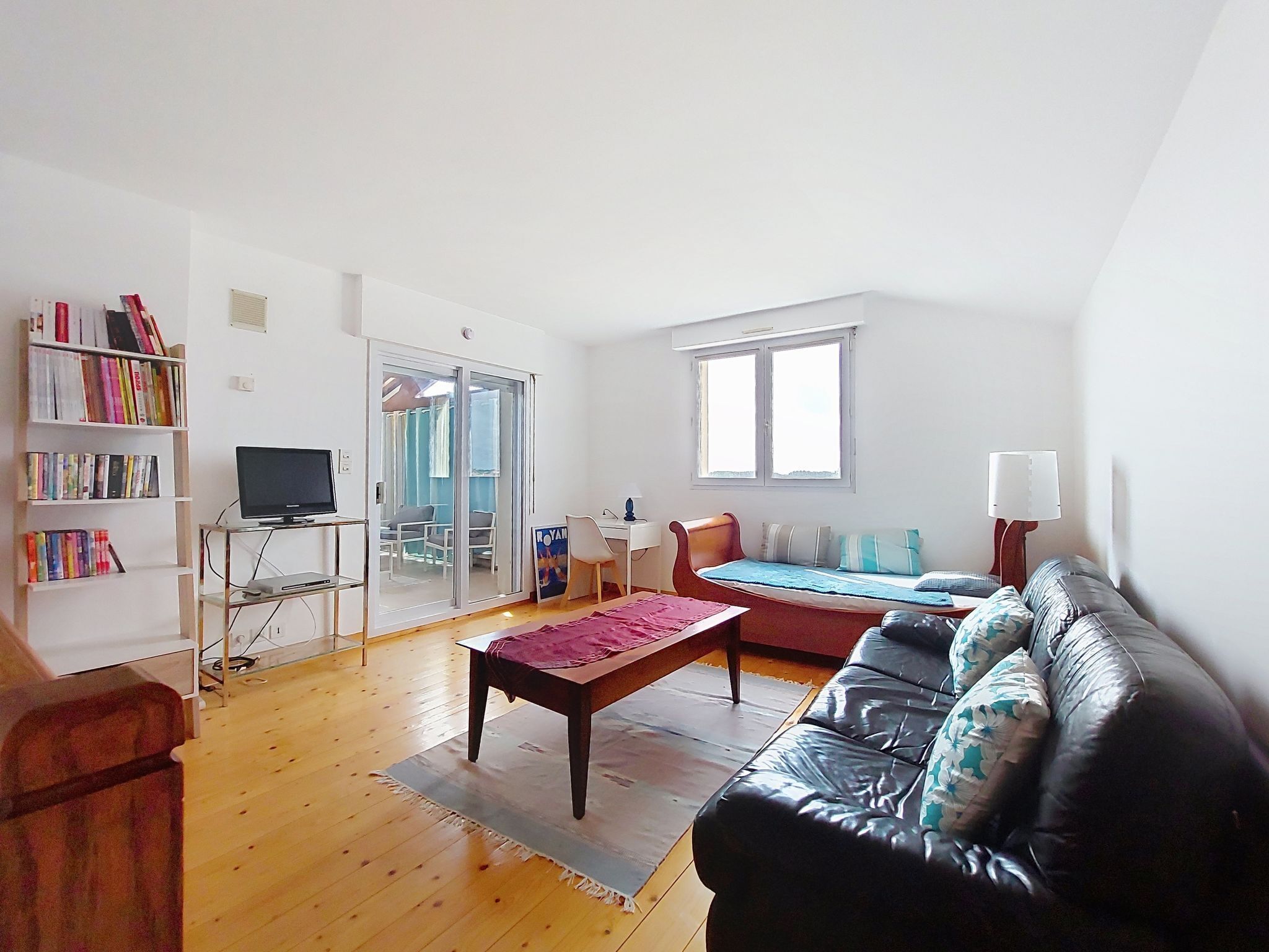 Een woonkamer met een zwarte leren bank, een houten salontafel, een boekenkast, een televisietoestel en een schuifdeur met glas die leidt naar een balkon.