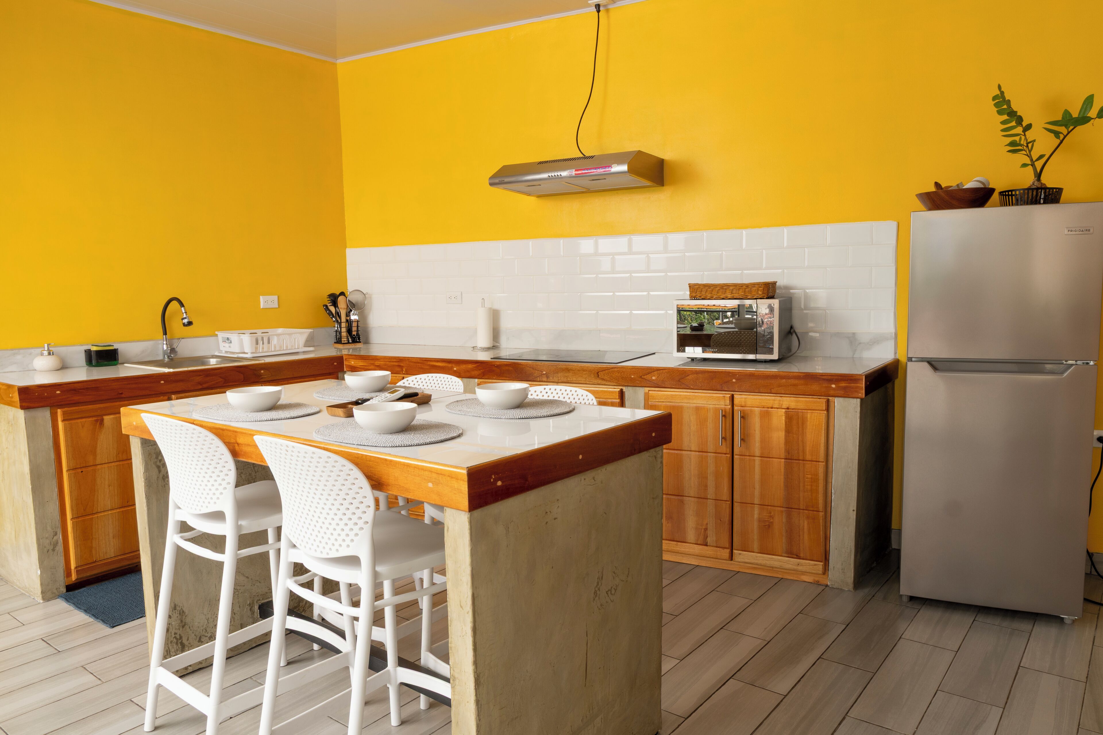 Una cocina con pared amarilla, salpicadero de azulejos blancos, armarios de madera, un refrigerador de acero inoxidable y una mesa de comedor de madera con sillas blancas.