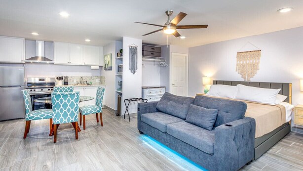 Une chambre d’hôtel moderne avec un canapé gris, un coin repas avec des chaises à motifs, une kitchenette et un ventilateur de plafond.
