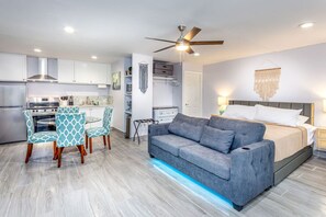 A modern hotel room with a grey sofa, a dining area with patterned chairs, a kitchenette, and a ceiling fan.