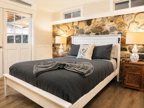 Una cama con un muro de piedra de fondo, una mesita de noche de madera y una puerta con ventanal.