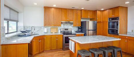 A modern kitchen with wooden cabinets, stainless steel appliances, a marble countertop, and a bar stool area.