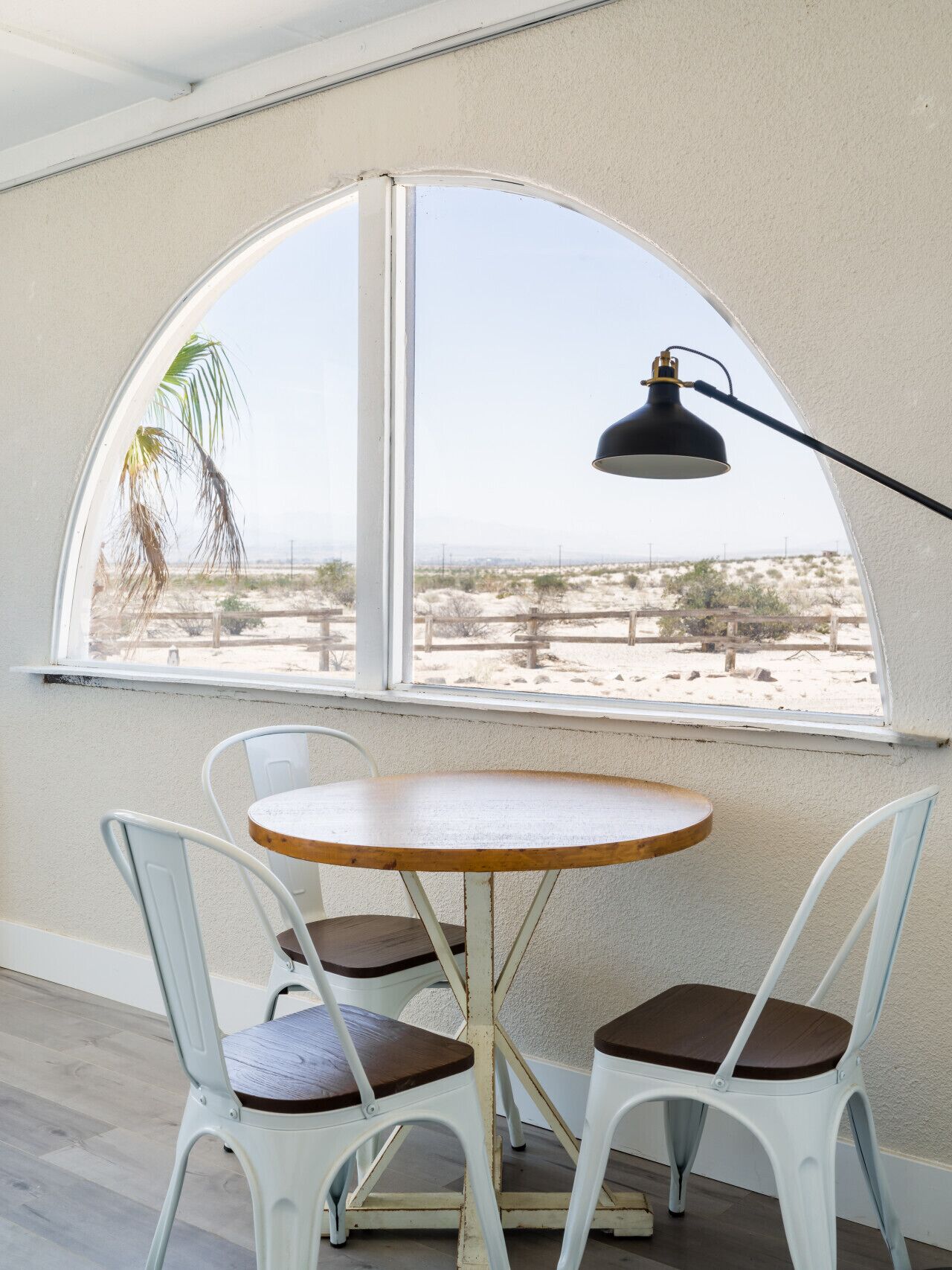 A room with a round table and two chairs, a window with a view of a desert landscape, and a black desk lamp.