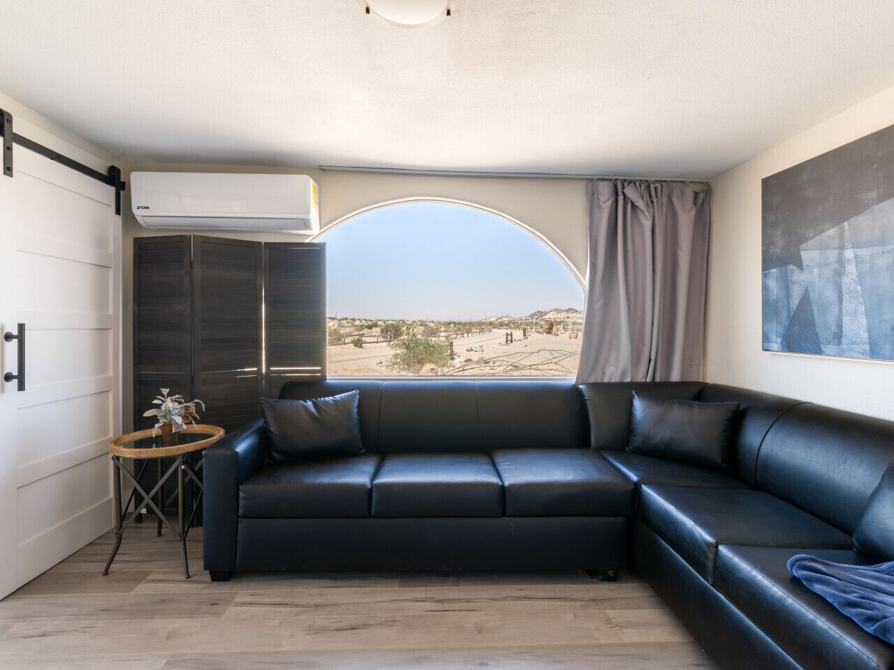 A modern living room with a black leather sofa, a small wooden side table, and a large window with a view of a desert landscape.