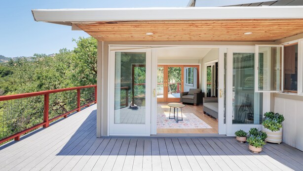 Una casa moderna con una veranda in legno, porte vetrate che conducono a un interno con un divano e un tavolo, e piante in vaso sulla veranda.