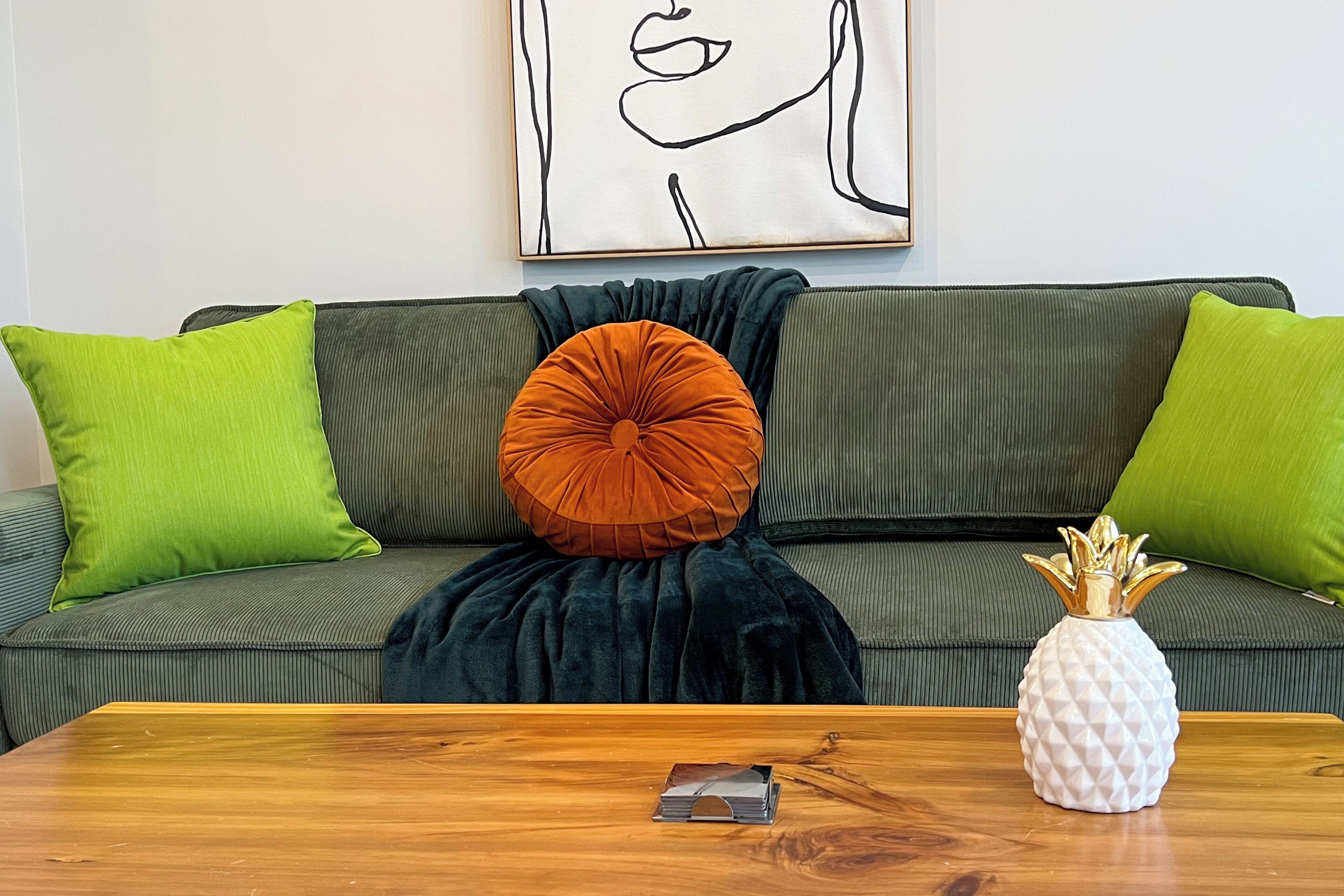 A green sofa with two green cushions, a dark blue throw blanket, and an orange cushion. A white pineapple-shaped vase is on a wooden table in front of the sofa.