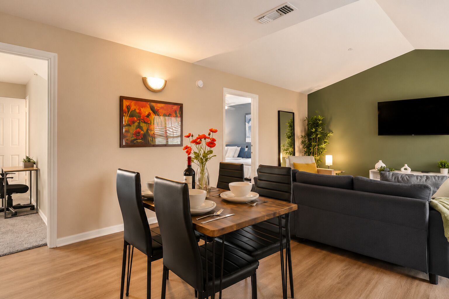 Una sala de estar moderna con comedor, un sofá, un televisor montado en la pared y una puerta que da a otra habitación.