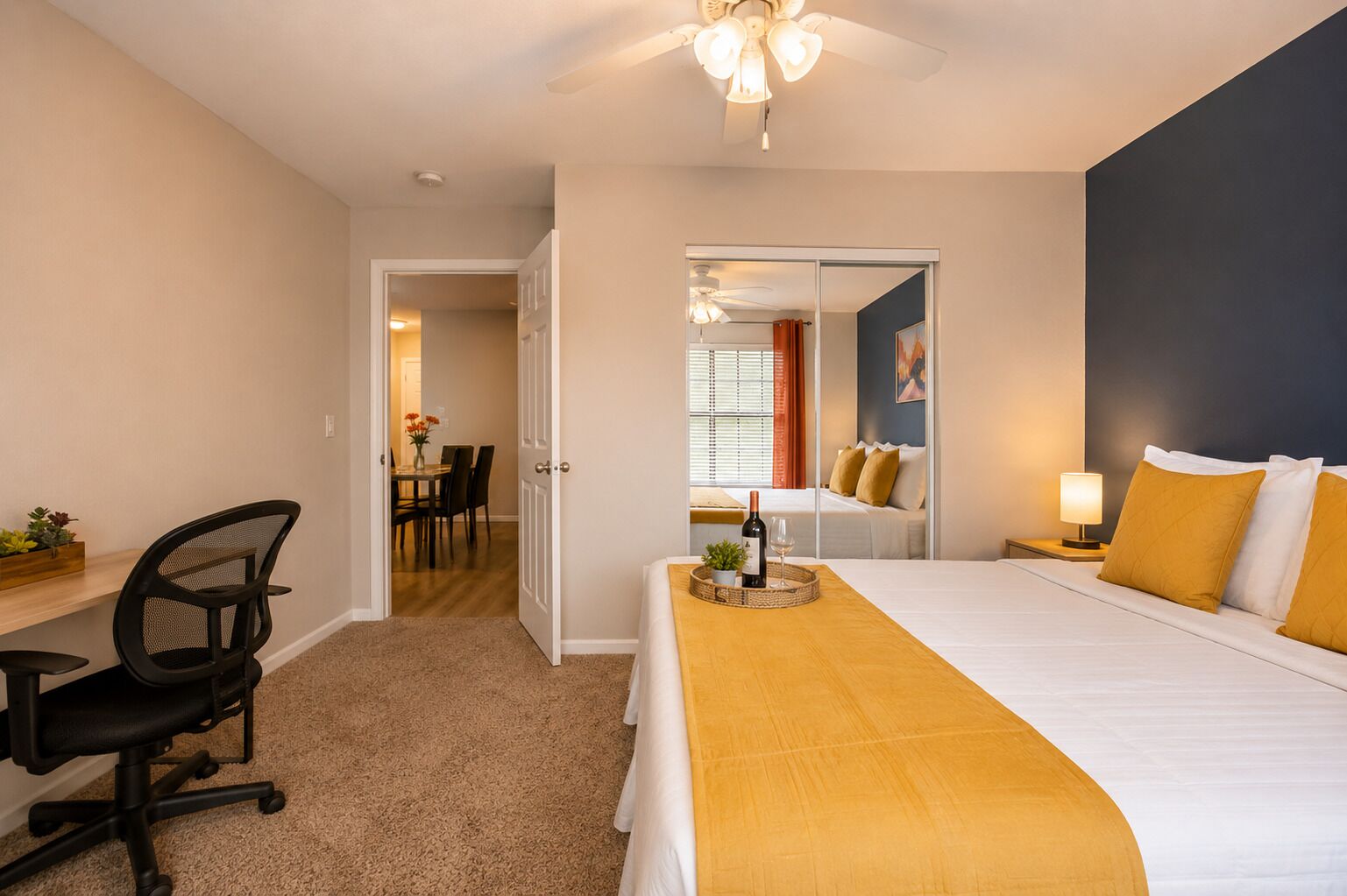 A hotel room with a bed, desk, chair, and a dining area visible through an open door.