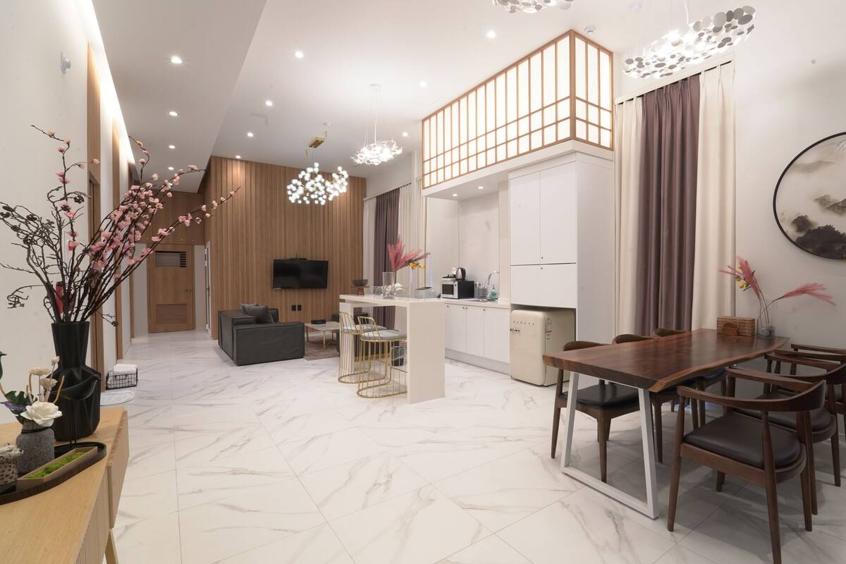 A modern living room with a sofa, dining table, and chairs, a kitchen area with a microwave, and a TV mounted on the wall.