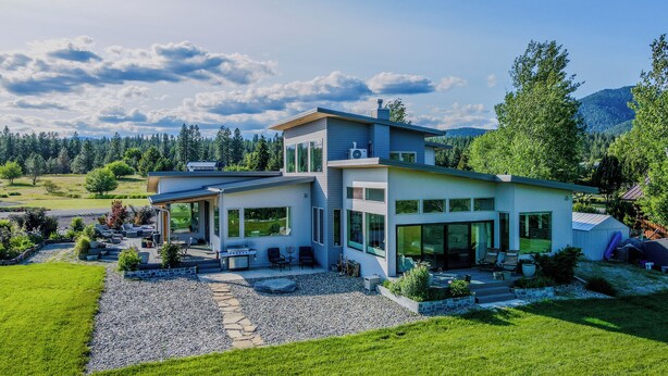 Une maison moderne à deux étages, dotée de grandes fenêtres, d’un patio avec mobilier d’extérieur et offrant une vue sur une zone boisée.