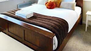 Een net opgemaakt bed met een bruin hoofdbord, wit beddengoed en een decoratief oranje kussen. Twee ingelijste illustraties van vogels hangen aan de muur, en er is een raam met een gestreept gordijn.