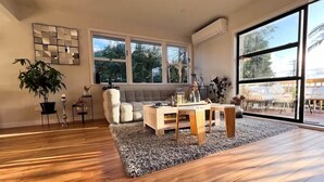 Een ruime woonkamer met een houten vloer, een grijs vloerkleed, een grijze bank en een houten salontafel. Er is een groot raam met uitzicht op bomen en een balkon.