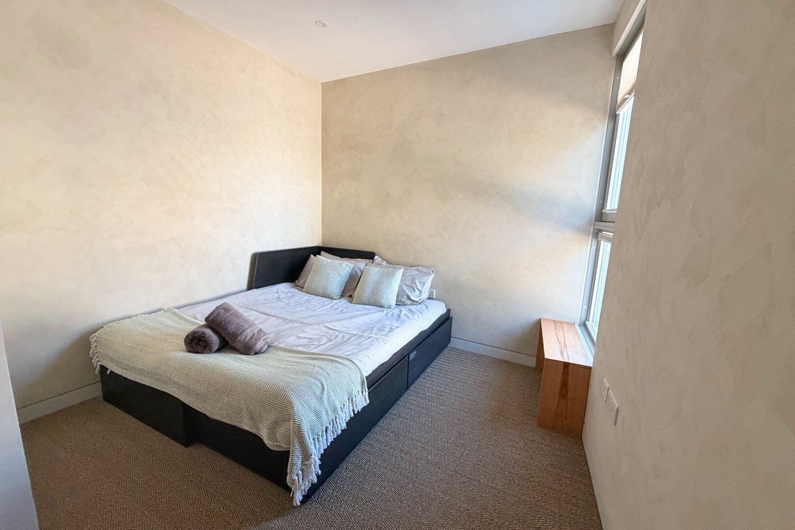 Een slaapkamer met een bed, kussens, een deken en een klein houten nachtkastje.