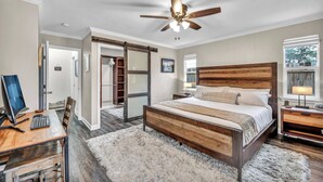 Une chambre à coucher avec un lit en bois, un bureau avec un ordinateur, un ventilateur de plafond et une grande fenêtre.