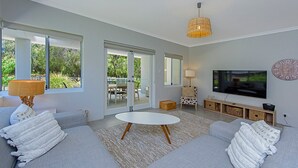 Una sala de estar moderna con un sofá gris, una mesa de centro blanca y un televisor en la pared. Hay un ventanal y una puerta que da a un espacio al aire libre.