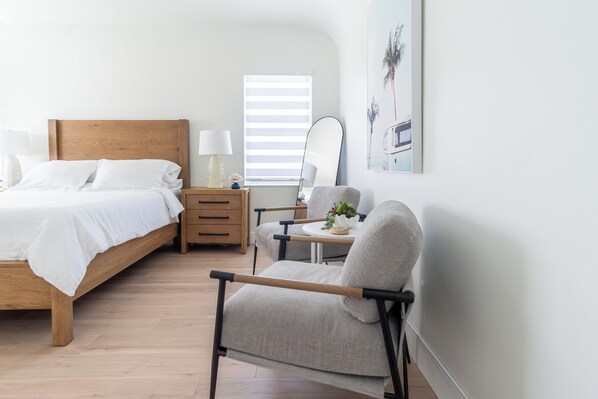 A bedroom with a wooden bed, a bedside table with a lamp, a chair, a small table with a plant, and a window with blinds. - Welcome to Sunrise & Palm Trees! (Miami Beach)