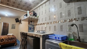 A compact kitchen with a built-in stove, a sink, and a small refrigerator. There is a water dispenser on the counter and a shelf above the stove holding kitchen items.