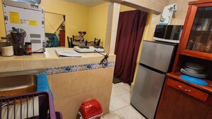 Eine kleine Küche mit Spüle, Herd, Mikrowelle und Kühlschrank. Ein Essbereich mit Stühlen und Tisch ist vorhanden, sowie ein kleines Fenster mit Vorhängen.