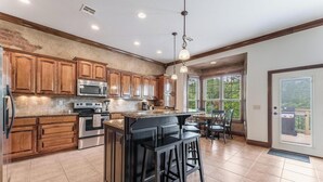 A spacious kitchen with wooden cabinets, a central island with bar stools, stainless steel appliances, and a view to the outside. - 5BR Bentonville Area Retreat | Sleeps 14 | 3 King Beds | GameRoom & Deck Firepit (Bella Vista)