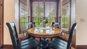 A round dining table with four chairs, centered in a bay window with blinds, overlooking a green outdoor area. - 5BR Bentonville Area Retreat | Sleeps 14 | 3 King Beds | GameRoom & Deck Firepit (Bella Vista)
