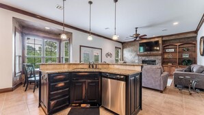 A modern kitchen with a large island, dark cabinetry, and a built-in dishwasher. There is a dining area with a table and chairs, and a seating area with a sofa and armchairs. - 5BR Bentonville Area Retreat | Sleeps 14 | 3 King Beds | GameRoom & Deck Firepit (Bella Vista)