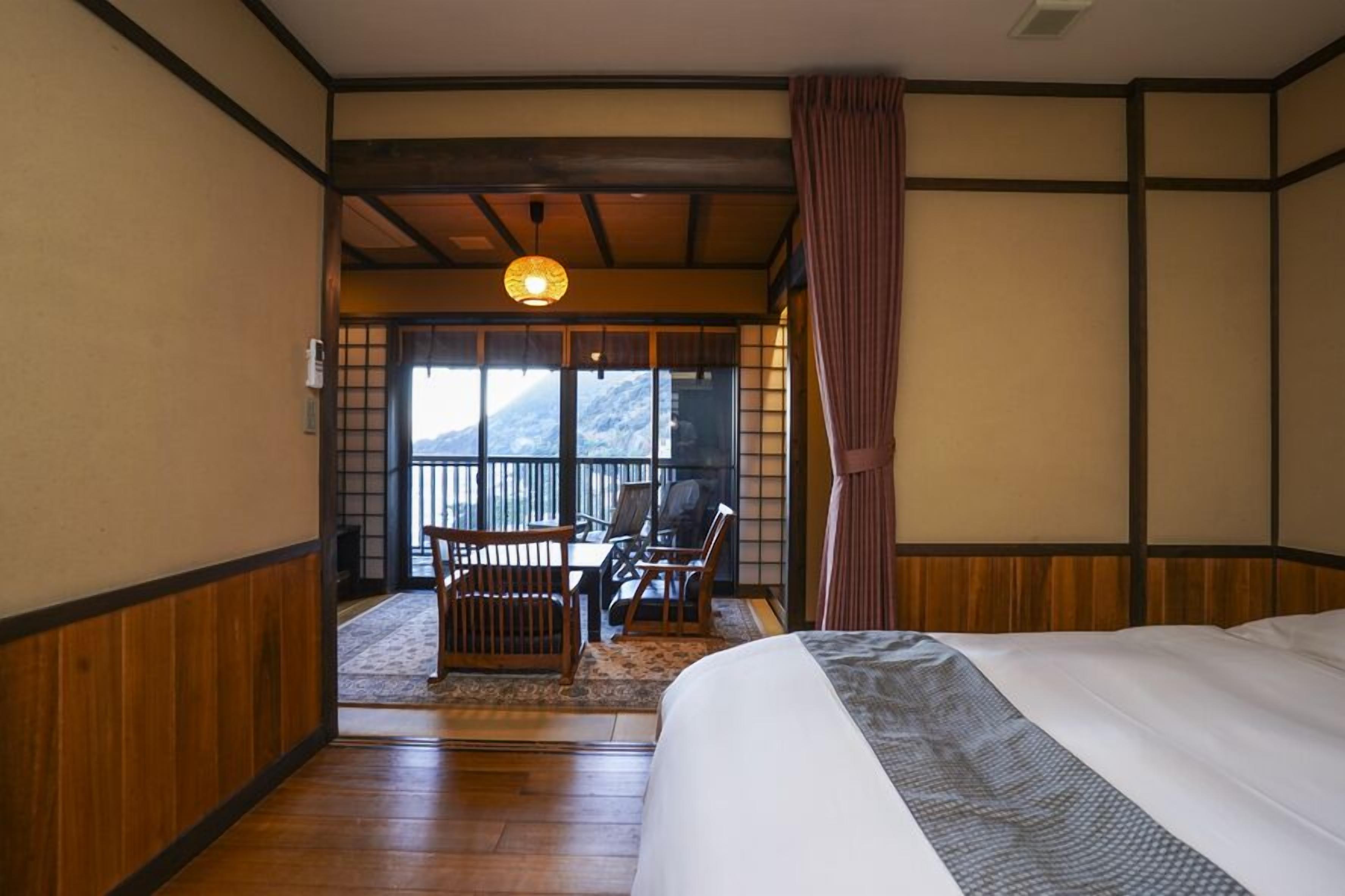 A traditional Japanese room with tatami flooring, a bed with a patterned bedspread, a wooden table with chairs, and a view of a mountain through a sliding door.