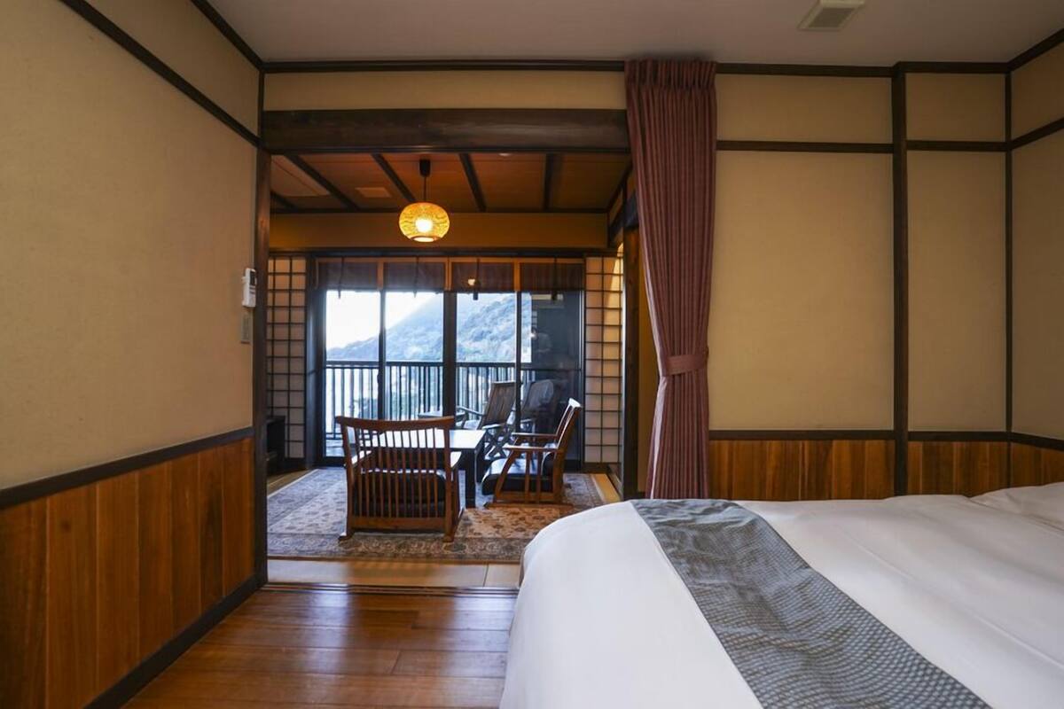 Una habitación japonesa tradicional con suelo de tatami, una cama con colcha estampada, una mesa de madera con sillas y vistas a la montaña a través de una puerta corredera.