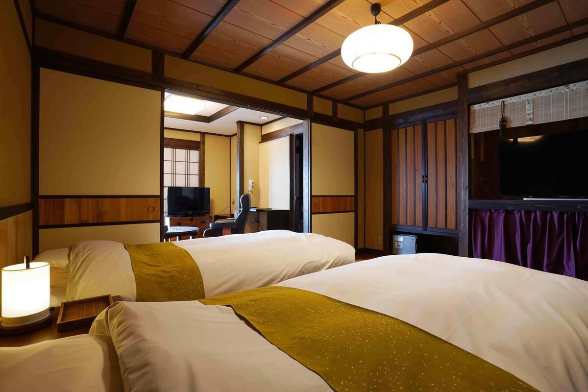 Habitación de hotel con dos camas, televisión, escritorio y baño visible a través de una puerta abierta.