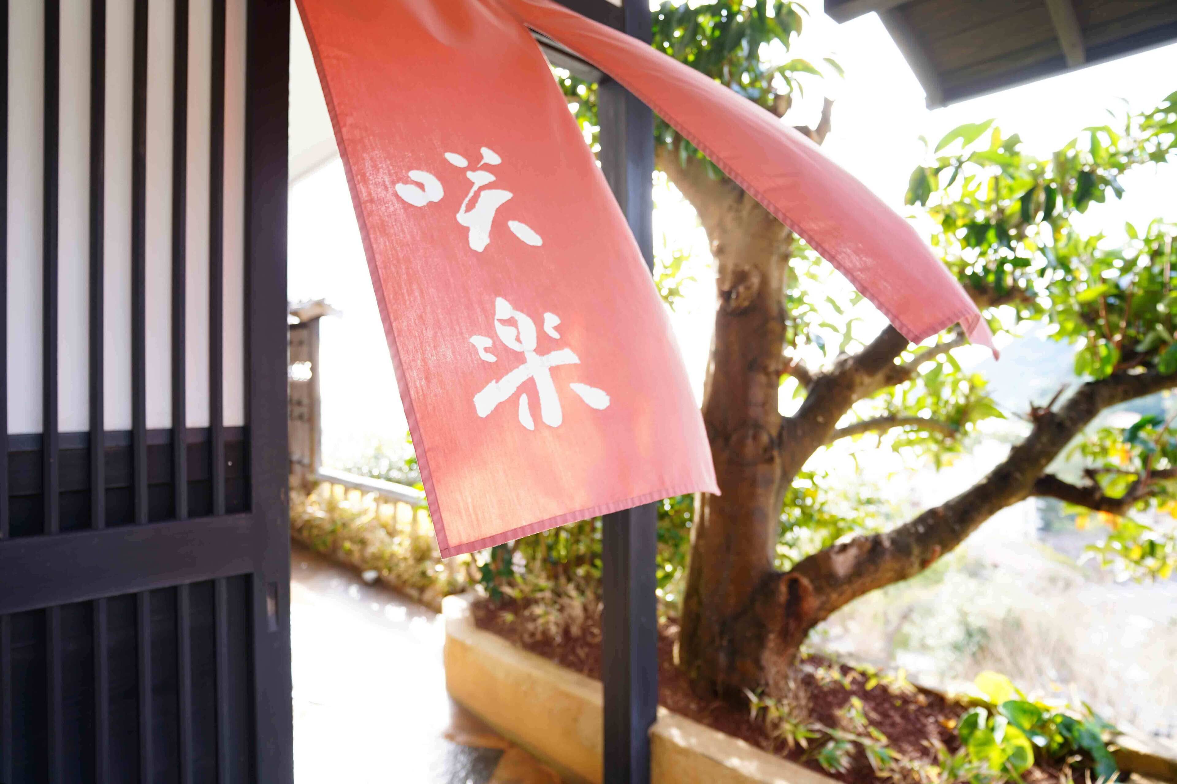 A red banner with white text hanging from a structure, a tree, and a building in the background.